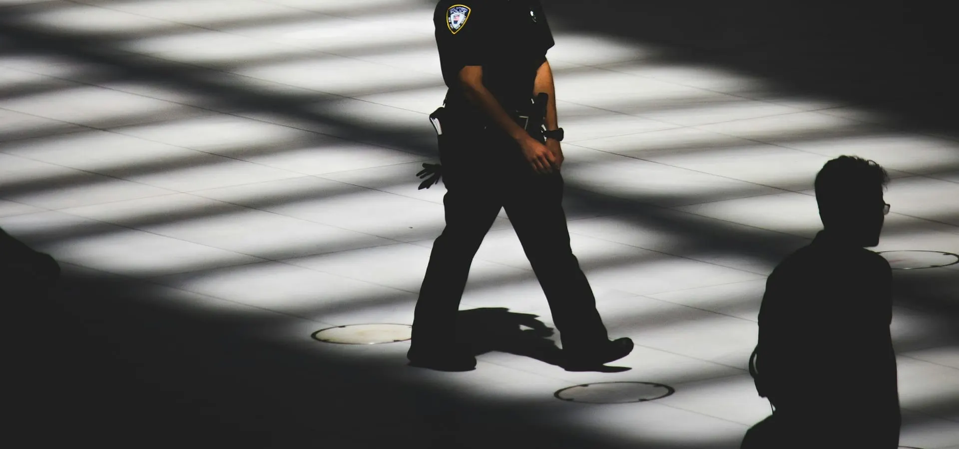 A uniformed security officer walking through bands of light and shadow inside a building.