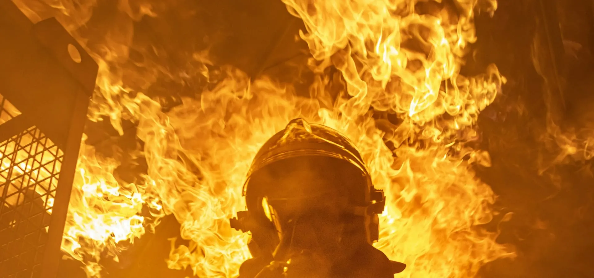 A firefighter in full turnout gear silhouetted against a large fire.